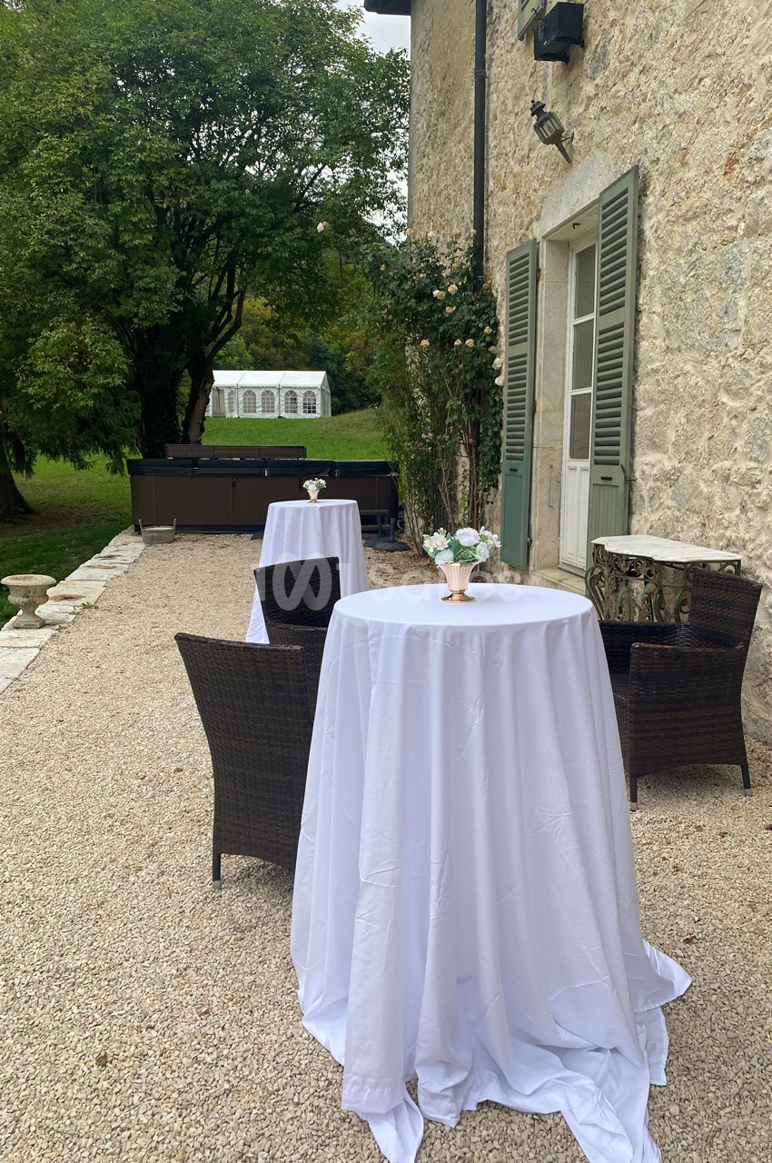 Table haute avec nappe blanche, entourée de chaises en osier, disposée sur une terrasse en gravier près d'un jardin.