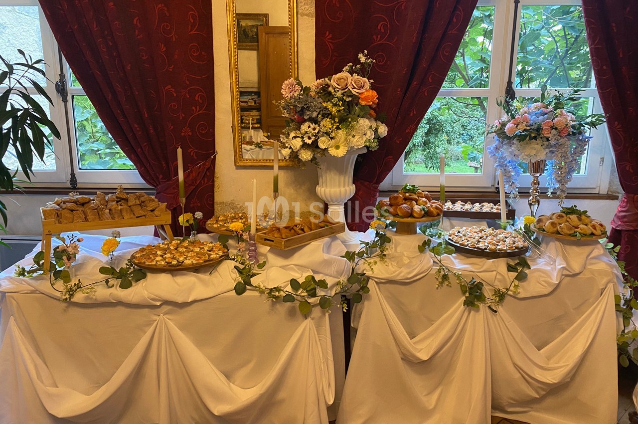 Table garnie de pâtisseries variées et décorée de fleurs, située devant des fenêtres avec rideaux rouges.