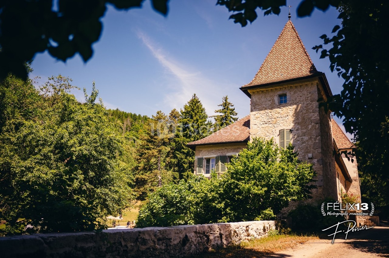 Maison en pierre avec toit en tuiles, entourée de verdure et d'arbres, sous un ciel bleu ensoleillé.