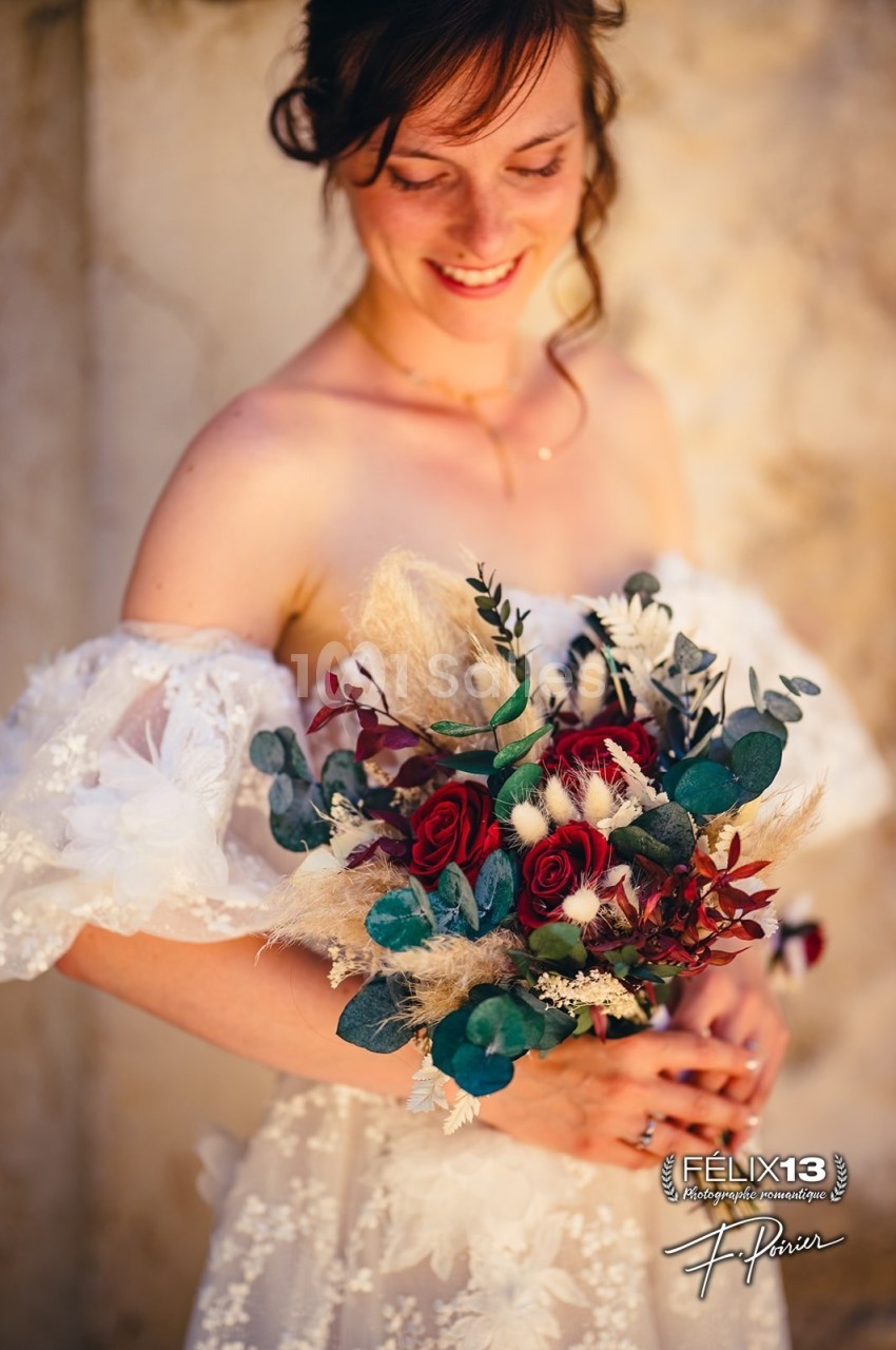 Femme en robe de mariée tenant un bouquet de fleurs rouges et blanches avec des feuillages, souriant devant un mur clair.