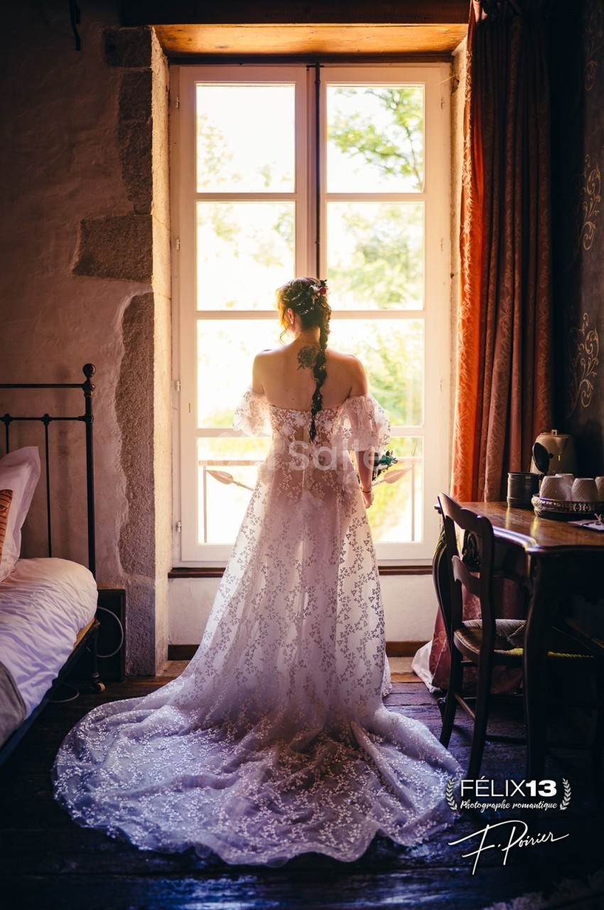 Une femme en robe de mariée en dentelle regarde par une fenêtre lumineuse dans une chambre au style rustique.