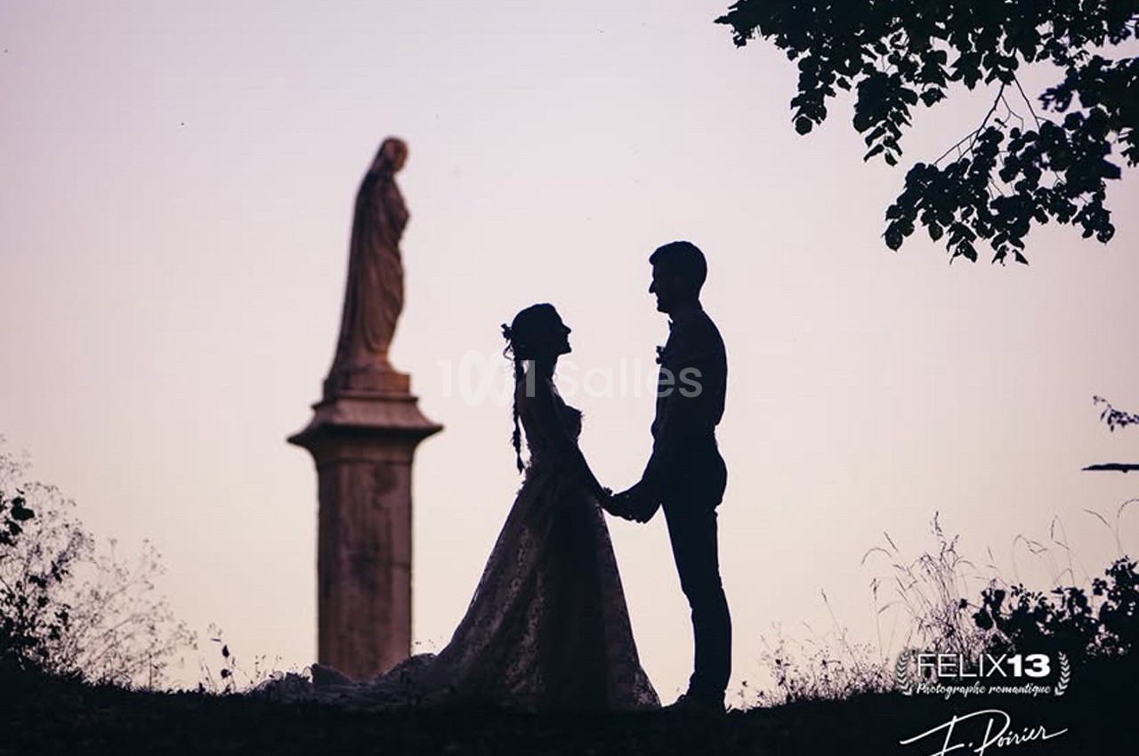 Un couple en tenue de mariage se tient la main devant une statue, au coucher du soleil.