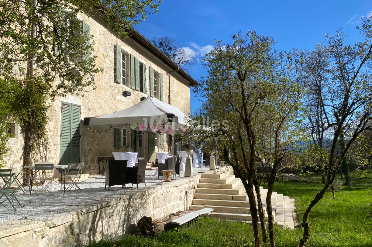 Terrasse d'une maison en pierre avec des tables dressées sous un barnum, entourée d'arbres et d'un jardin verdoyant.