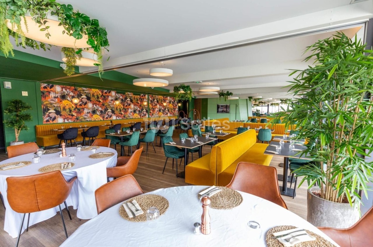 Salle de restaurant lumineuse avec tables dressées, banquettes jaunes, plantes décoratives et fresque colorée au mur.