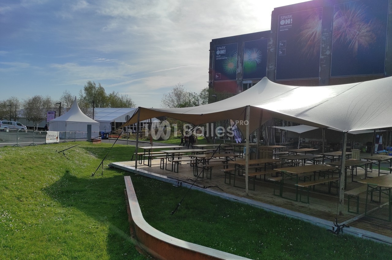 Terrasse extérieure avec tables et bancs sous des tentes, située près d'un bâtiment et entourée d'espaces verts.