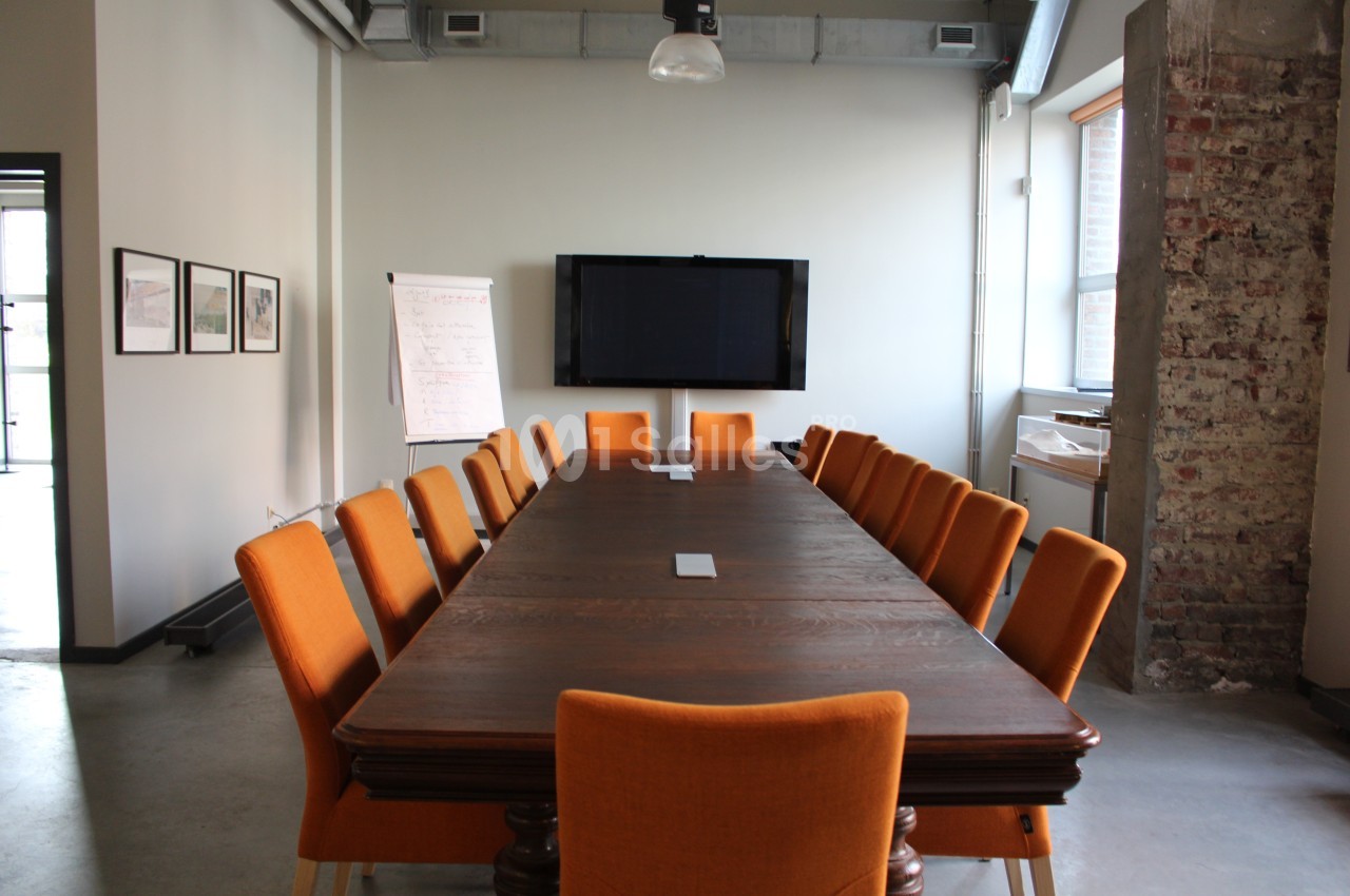 Salle de réunion avec une grande table en bois entourée de chaises orange, un écran mural et un tableau de conférence.