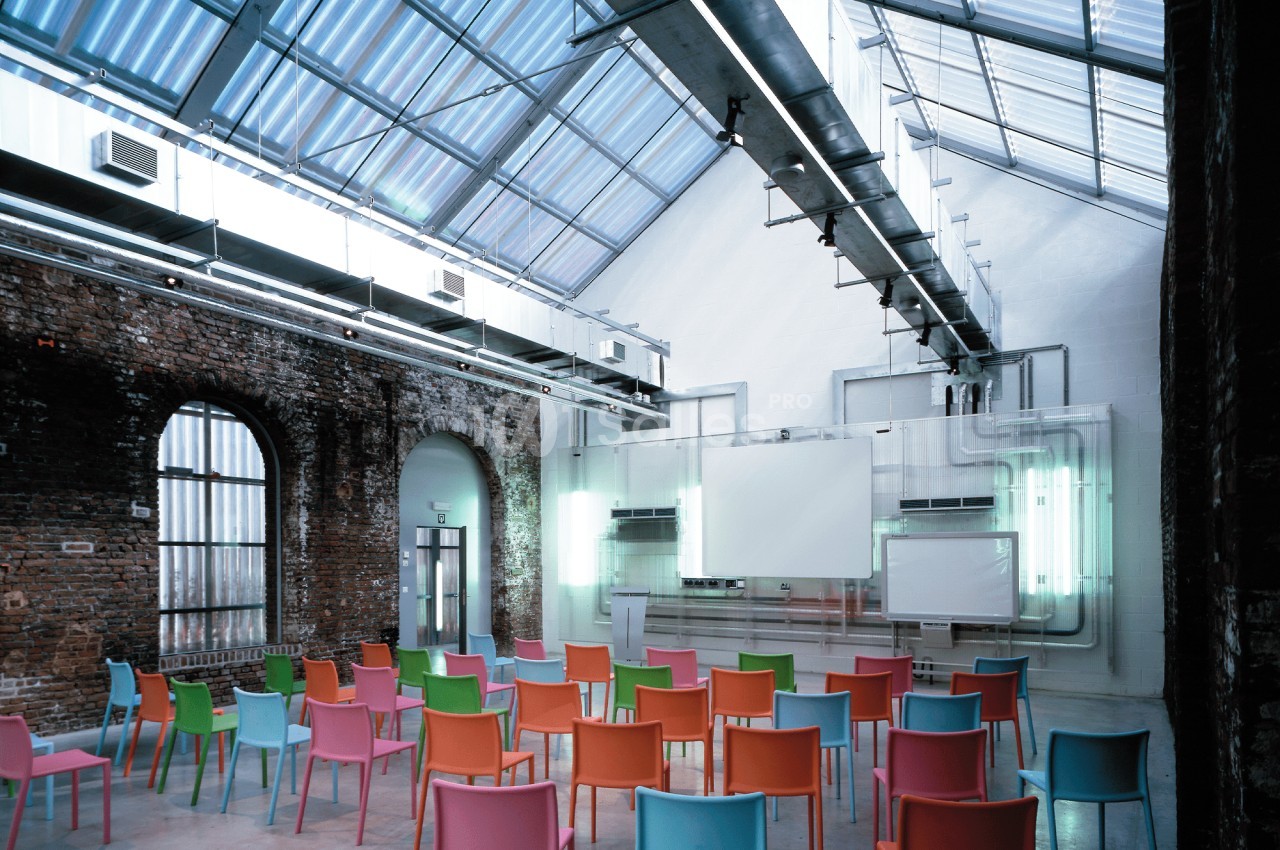 Salle de conférence lumineuse avec des murs en briques, des chaises colorées et des écrans de projection.