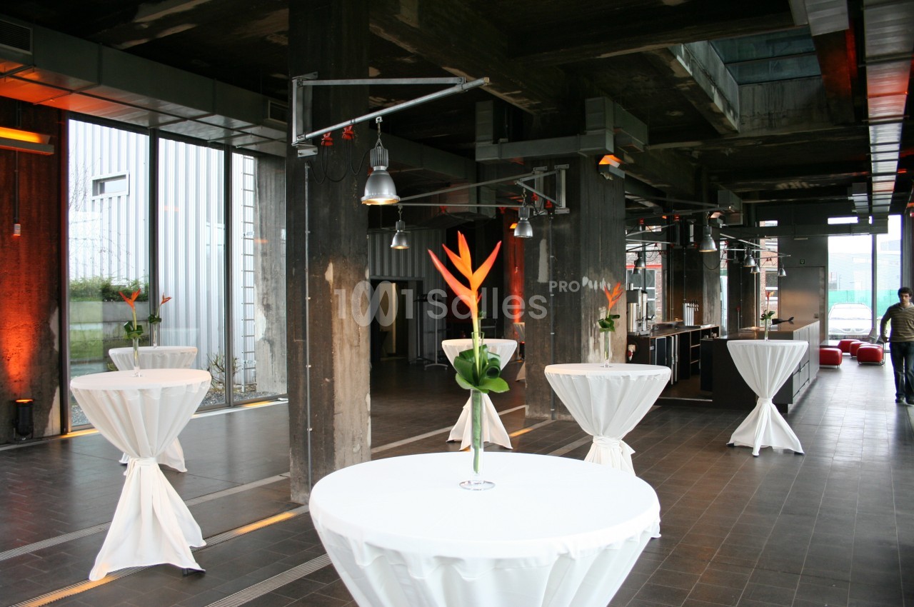 Salle moderne avec tables hautes drapées de blanc, décorées de fleurs tropicales, éclairage industriel.