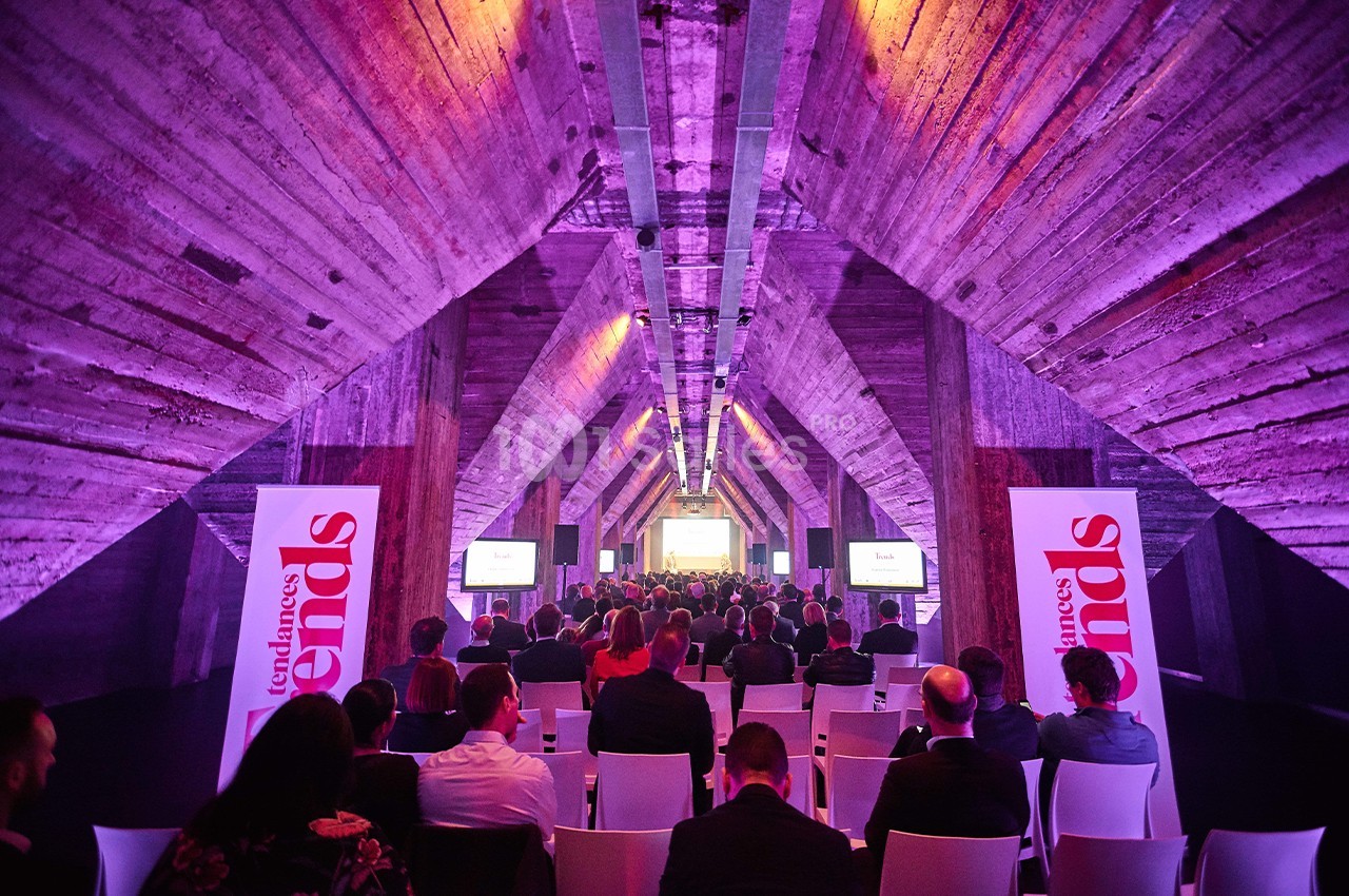 Salle de conférence avec un public assis, éclairage violet et écrans diffusant une présentation.
