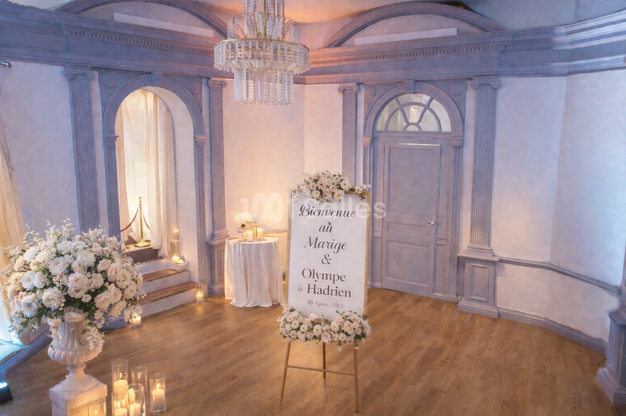 Salle décorée pour un mariage avec un panneau de bienvenue entouré de fleurs et des bougies disposées au sol.