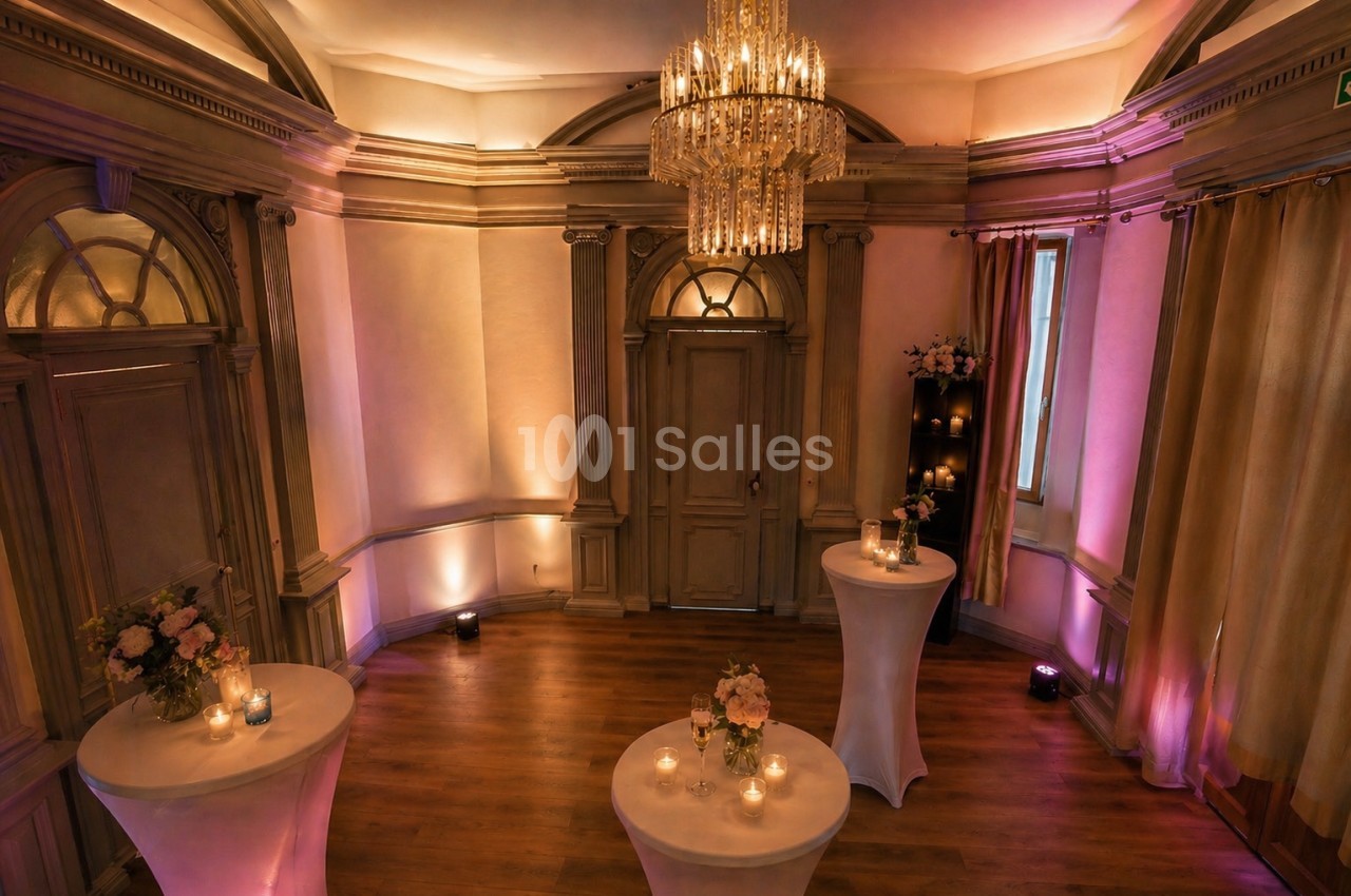 Salle élégante avec parquet, tables hautes décorées de fleurs et bougies, éclairage tamisé et lustre central.