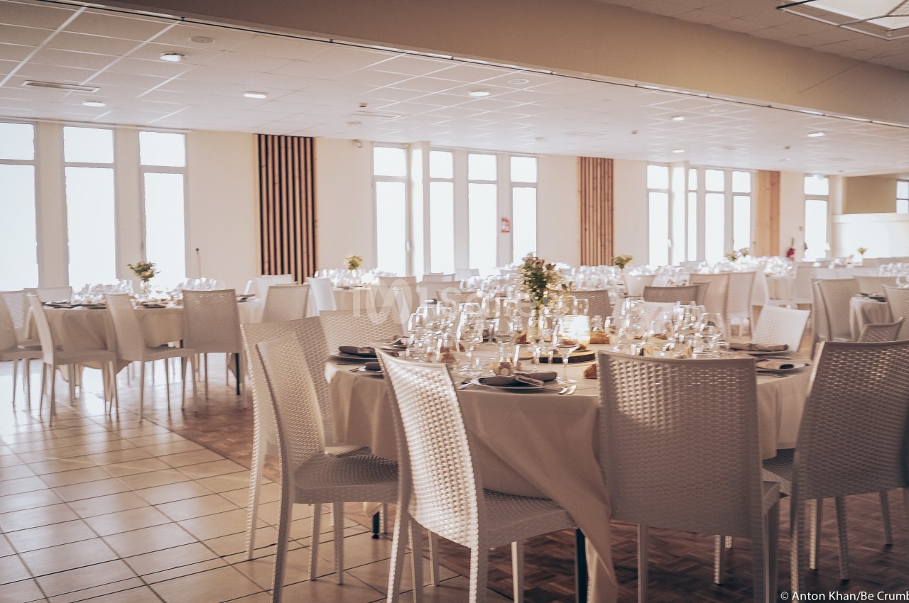 Salle de réception lumineuse avec tables rondes dressées, nappes blanches et chaises en rotin blanc.