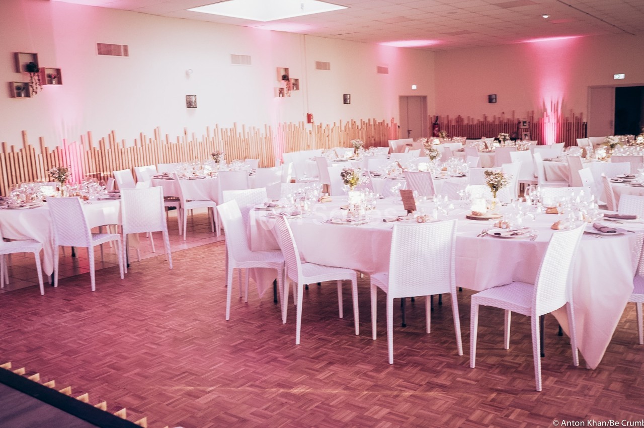 Salle de réception décorée avec des tables rondes et carrées, nappes blanches et éclairage rose tamisé.