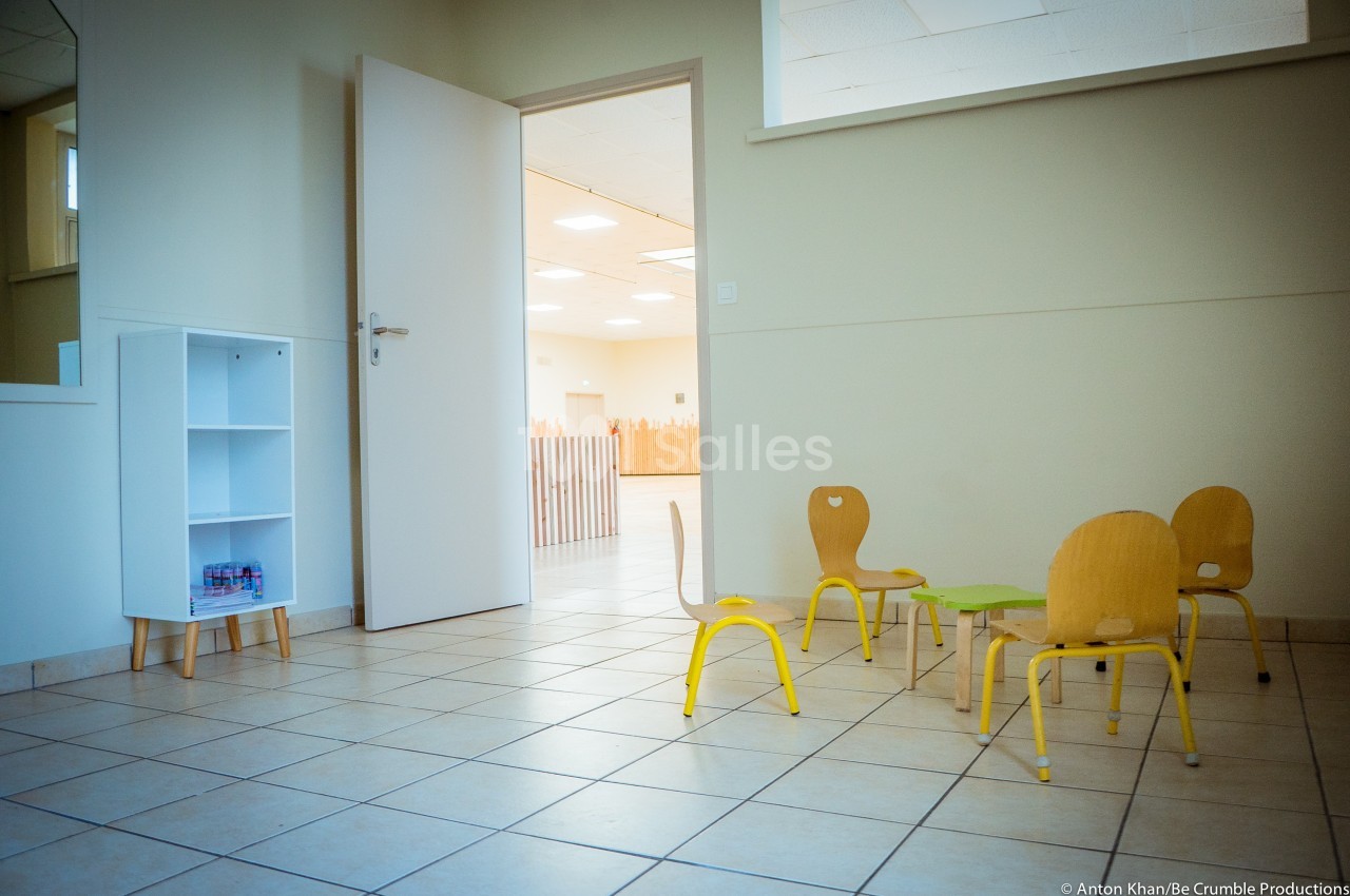 Salle lumineuse avec chaises d'enfants en bois, petite table verte et étagère blanche près d'une porte ouverte.