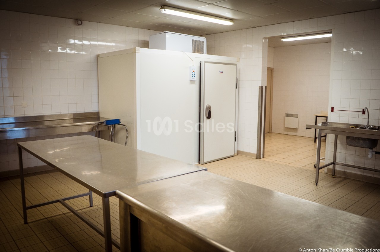 Cuisine professionnelle avec plans de travail en inox, carrelage blanc et une grande chambre froide au fond.