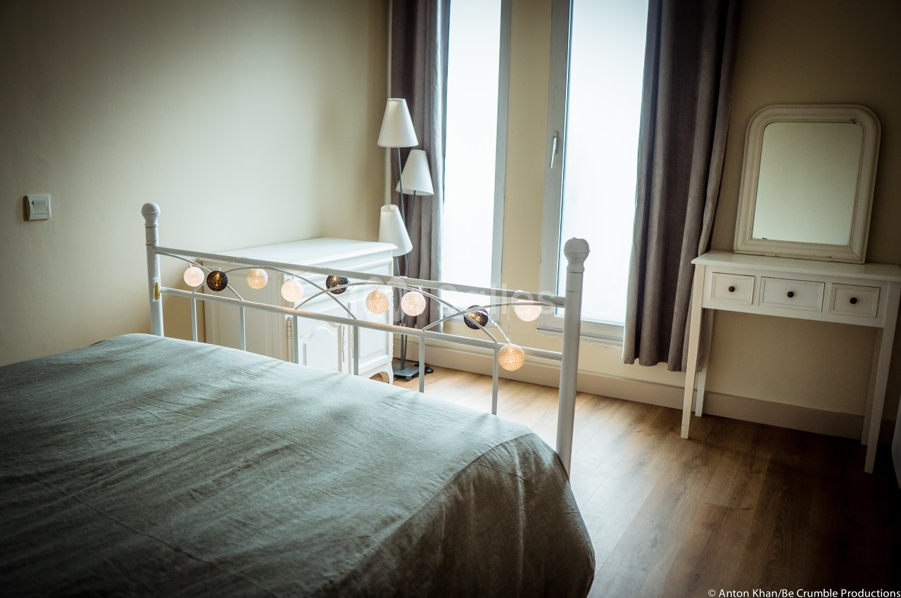 Chambre lumineuse avec lit à cadre métallique, guirlande lumineuse, bureau blanc et grande fenêtre avec rideaux.