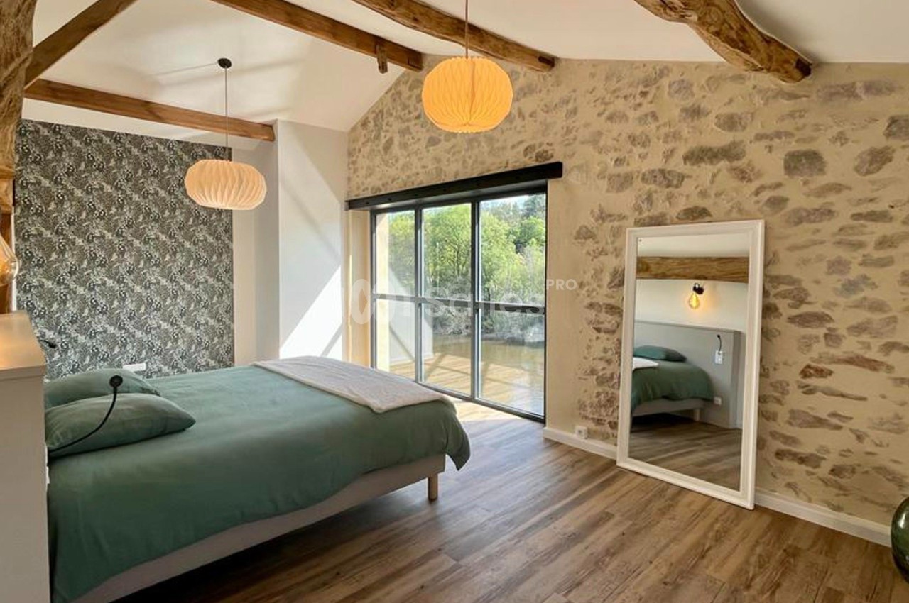 Chambre lumineuse avec lit double, mur en pierre, parquet, miroir sur pied et grande baie vitrée donnant sur l'extérieur.