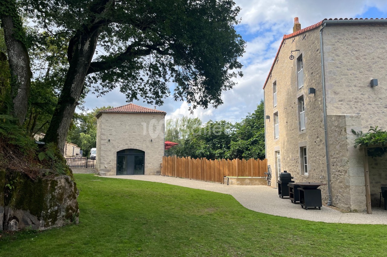 Cour avec pelouse, bâtiment en pierre, clôture en bois et arbres sous un ciel partiellement nuageux.