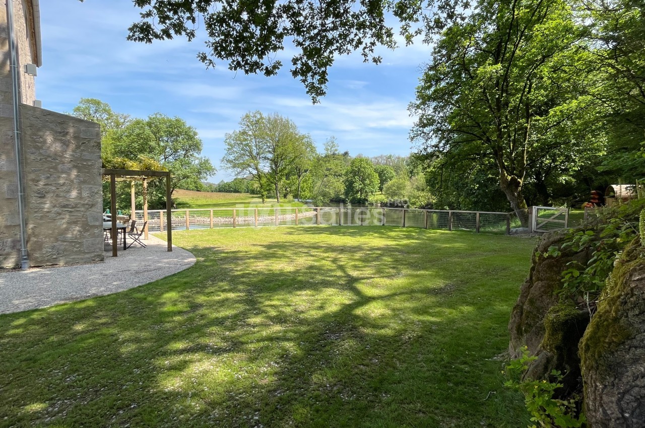 Jardin verdoyant avec pelouse, arbres et clôture en bois, bordé par une terrasse en pierre sous un ciel dégagé.