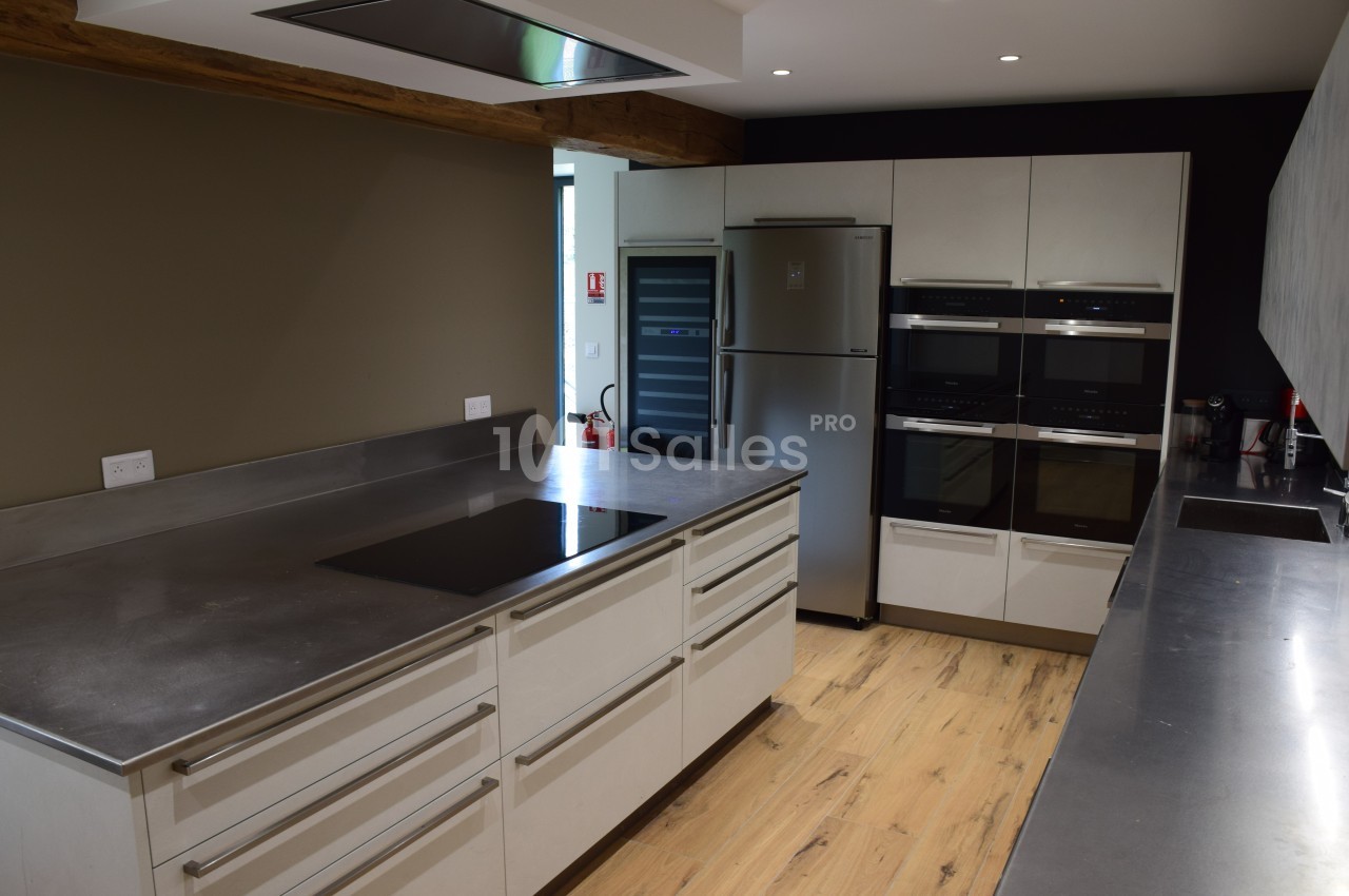 Cuisine moderne avec plan de travail en inox, plaques de cuisson, réfrigérateur, fours encastrés et sol en bois clair.