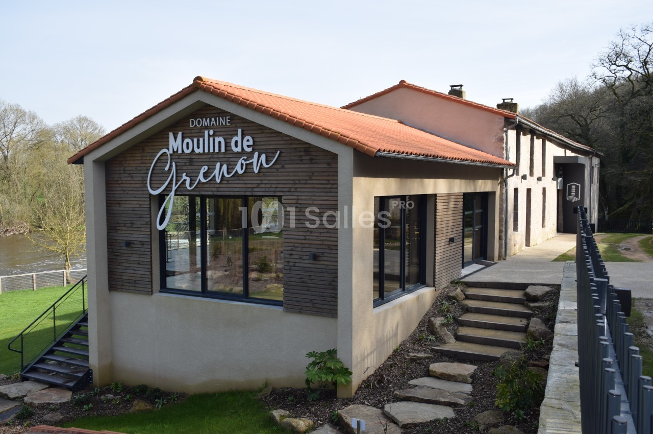 Bâtiment moderne avec façade en bois et inscription ’Moulin de Grenon’, situé près d'une rivière et entouré de verdure.