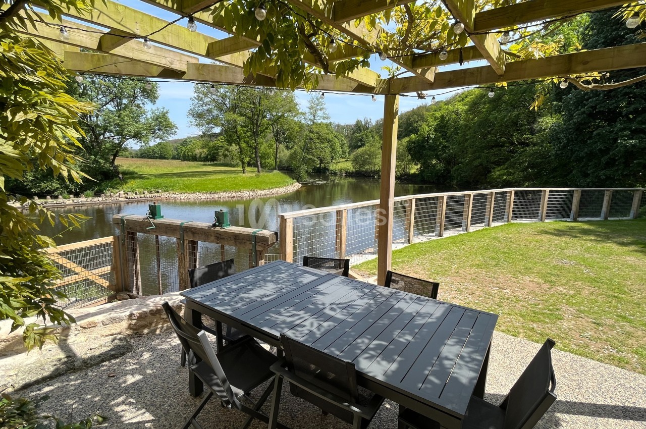 Table et chaises noires sous une pergola en bois, avec vue sur un étang entouré de verdure et une barrière en bois.
