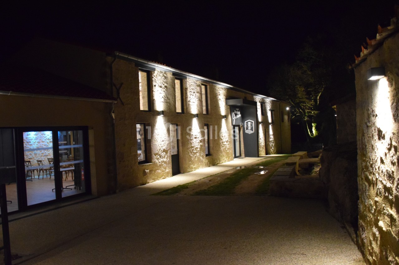 Façade en pierre d'un bâtiment éclairé la nuit, avec une entrée principale et une salle visible à travers une baie vitrée.