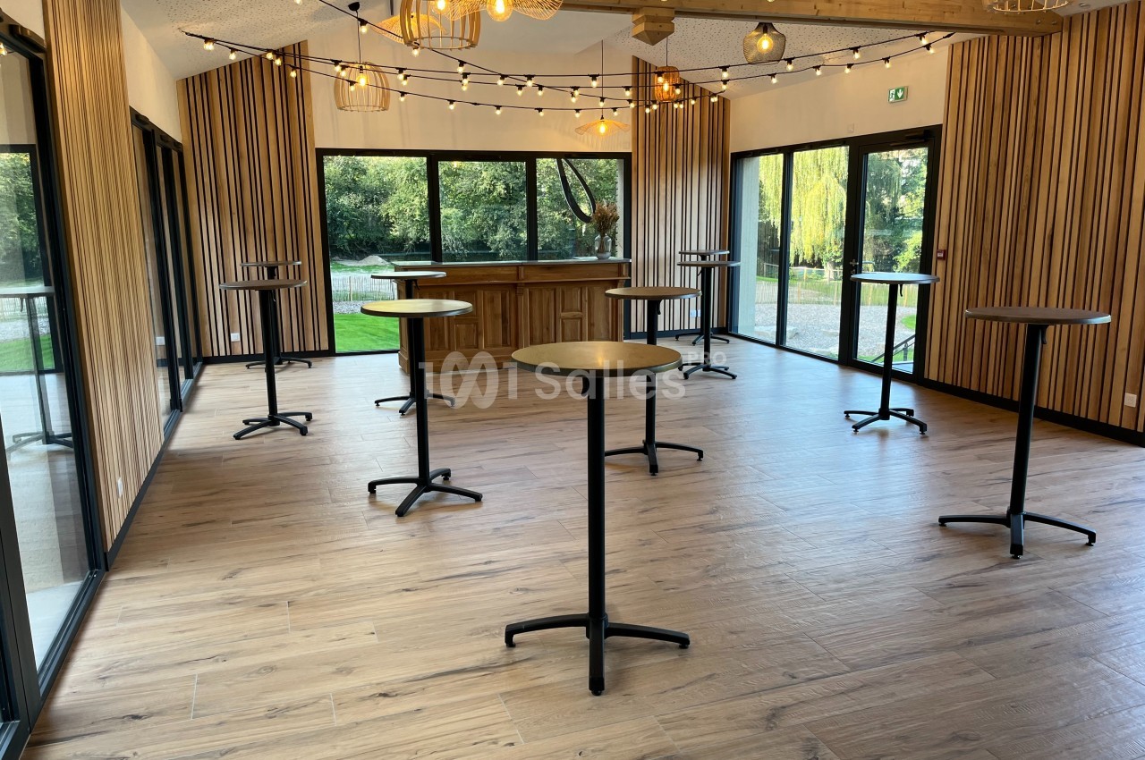 Salle lumineuse avec parquet, tables hautes noires, murs en bois et grandes baies vitrées donnant sur un jardin.