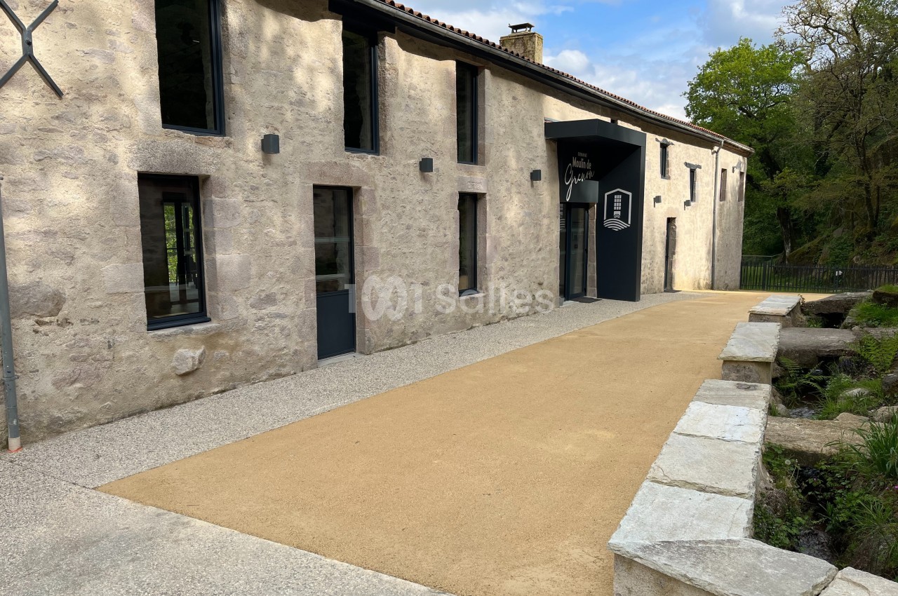 Bâtiment en pierre rénové avec entrée moderne, chemin en gravier et végétation en arrière-plan.