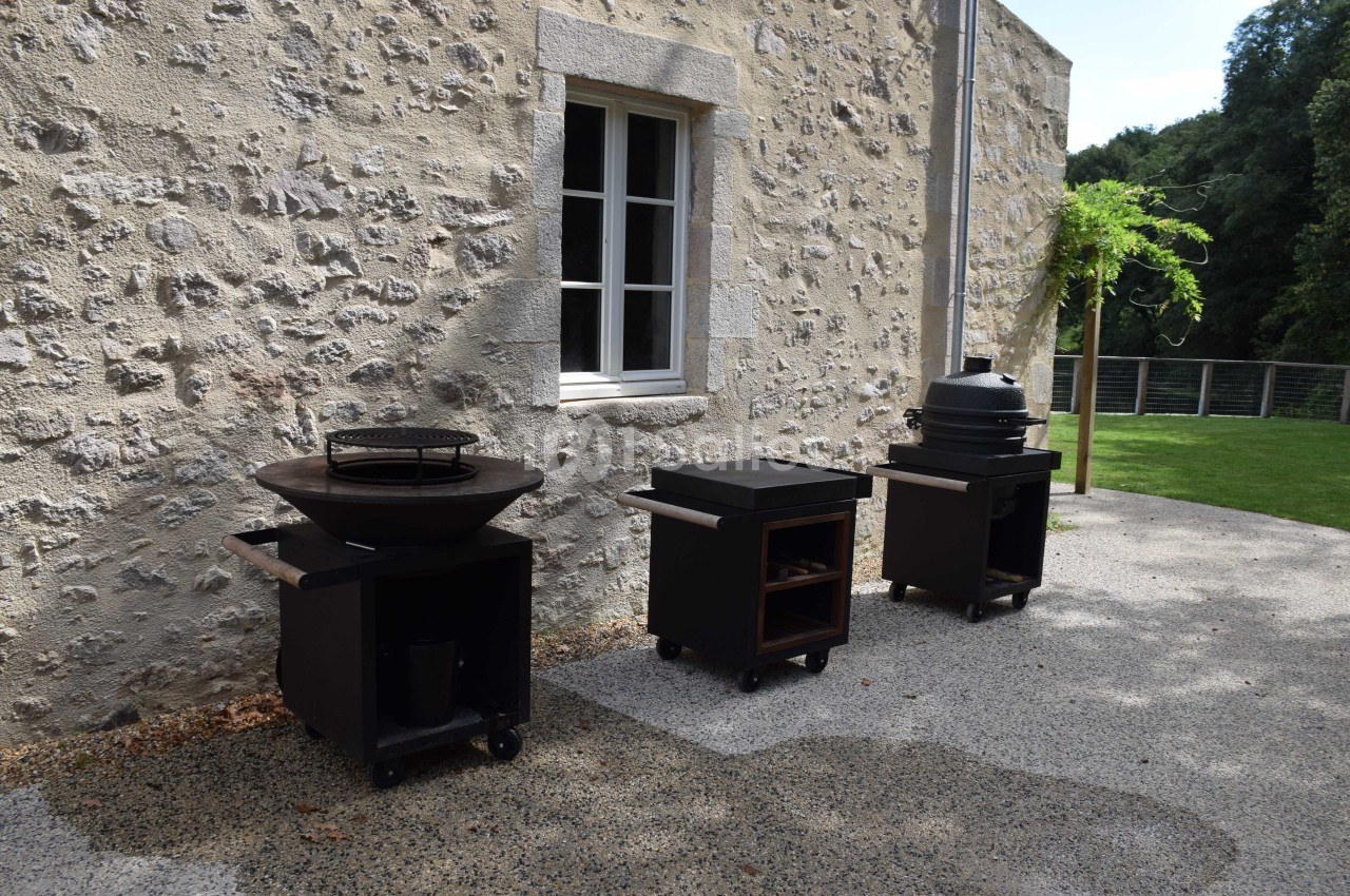 Trois barbecues modernes sur roulettes alignés devant un mur en pierre, près d'une fenêtre et d'un espace vert.