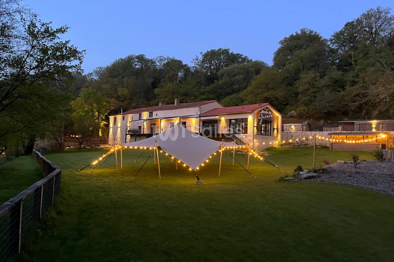 Bâtiment en pierre éclairé au crépuscule, avec une tente lumineuse installée sur une pelouse verdoyante.