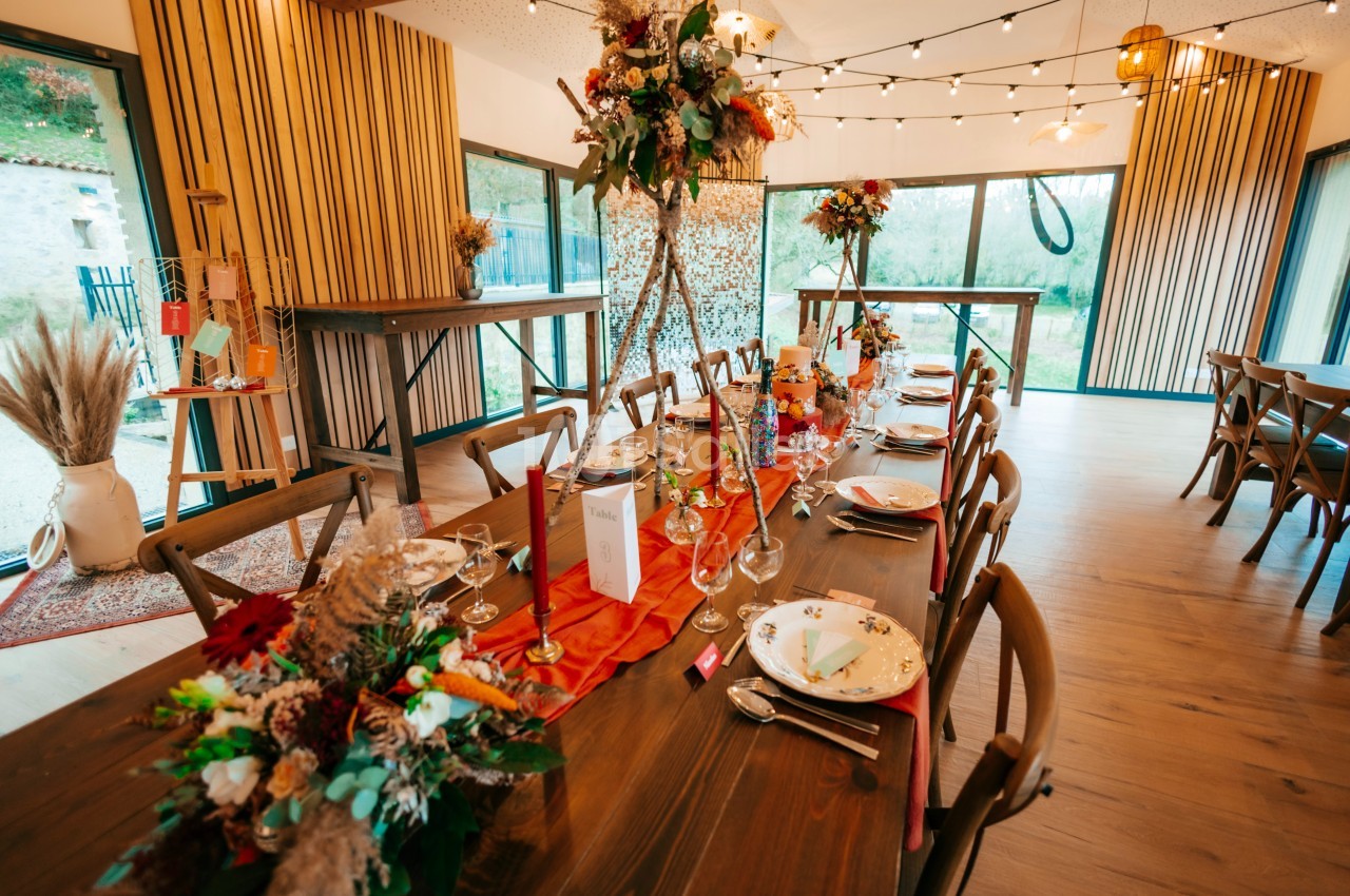 Table en bois décorée pour un événement, avec fleurs, vaisselle élégante et guirlandes lumineuses en arrière-plan.