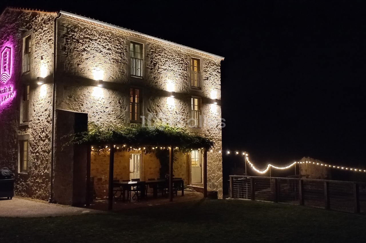 Bâtiment en pierre éclairé de nuit avec une enseigne lumineuse rose et une terrasse décorée de guirlandes.