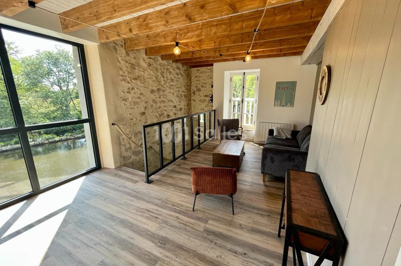 Espace lumineux avec parquet, murs en pierre, poutres apparentes, baies vitrées et mobilier moderne, vue sur un cours d'eau.