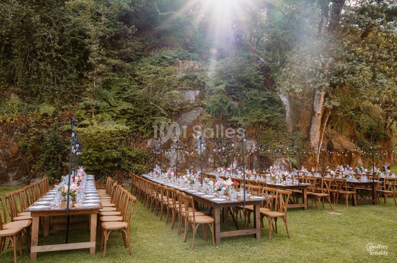 Tables dressées pour un repas en plein air dans un cadre naturel, entourées de végétation et éclairées par le soleil.