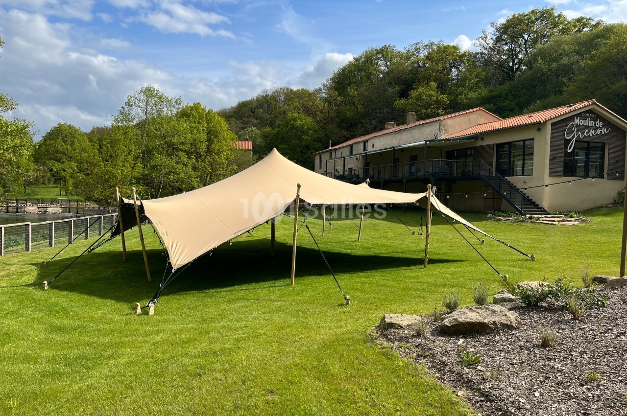 Tente beige montée sur une pelouse verte devant un bâtiment en pierre entouré de végétation.