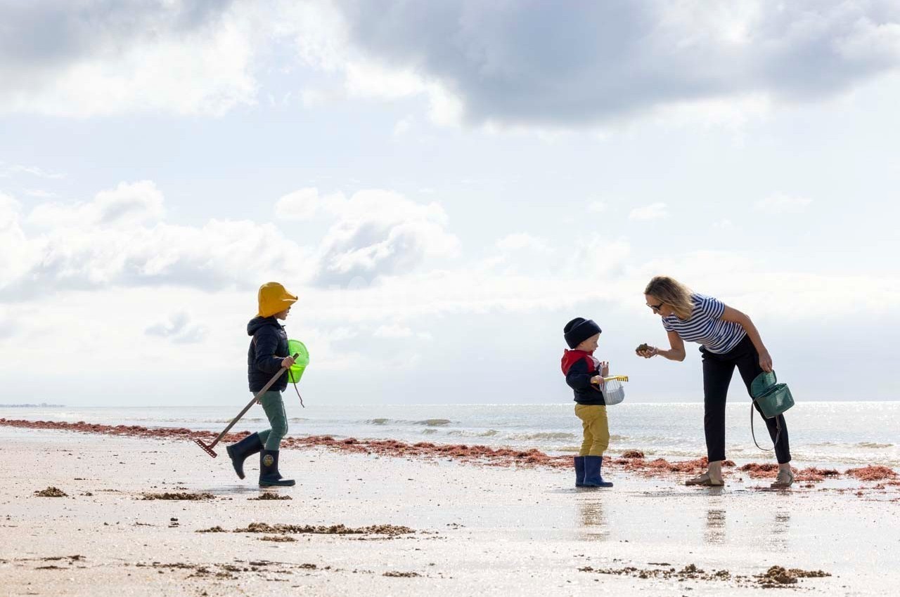 Une femme interagit avec un enfant sur une plage, tandis qu'un autre enfant marche avec une pelle et un seau.