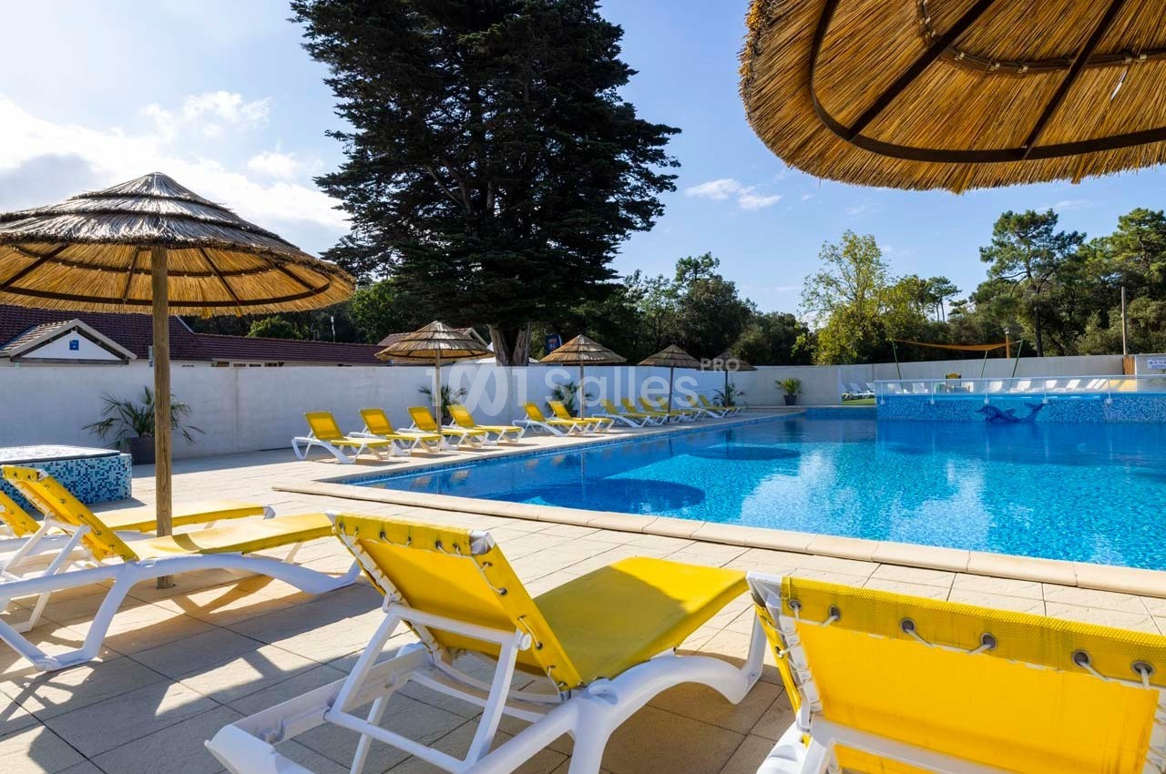 Chaises longues jaunes et parasols en paille disposés autour d'une piscine extérieure sous un ciel dégagé.