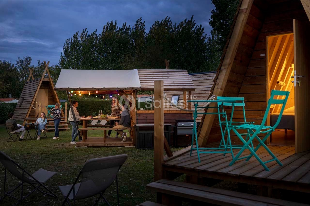 Des personnes dînent sous un auvent éclairé près de cabanes en bois triangulaires, entourées de mobilier de jardin.