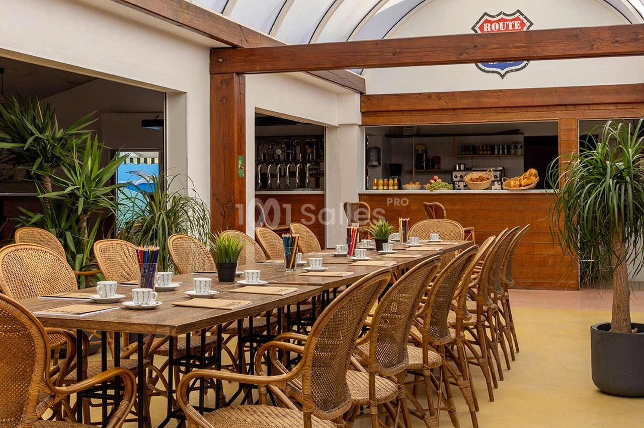 Salle lumineuse avec de grandes tables en bois, chaises en rotin, plantes vertes et un comptoir en arrière-plan.