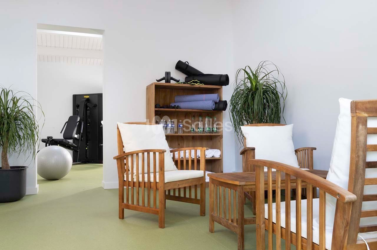 Espace détente avec fauteuils en bois, plantes et étagère contenant des accessoires de sport, vue sur une salle de fitness.