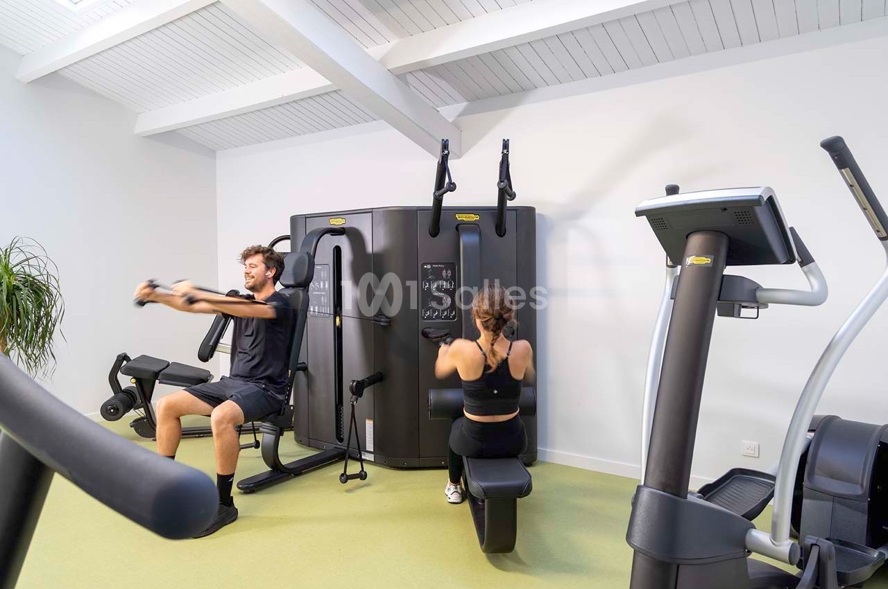 Un homme et une femme utilisent des équipements de musculation dans une salle de sport lumineuse.