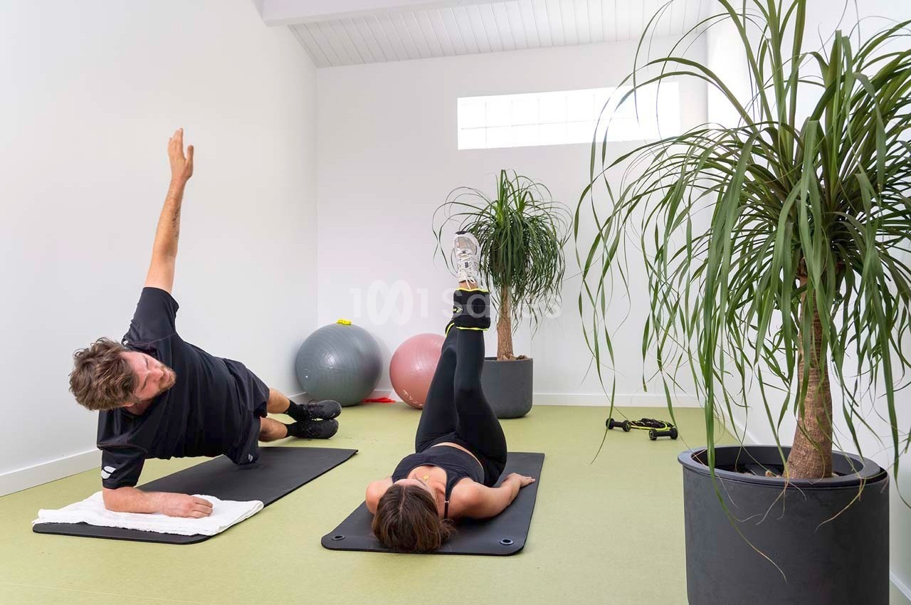 Deux personnes réalisant des exercices de renforcement musculaire sur des tapis dans une salle lumineuse avec des plantes.