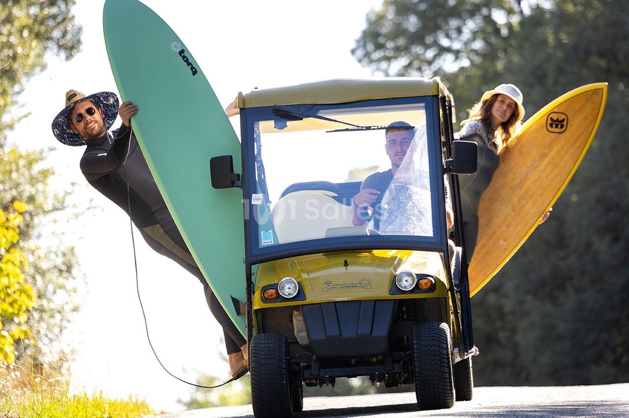Trois personnes en combinaison de surf transportant des planches autour d'une petite voiturette jaune sur une route…