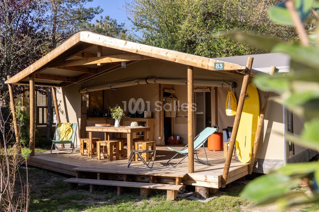 Tente lodge en bois avec terrasse aménagée, mobilier extérieur et équipements, entourée de verdure.