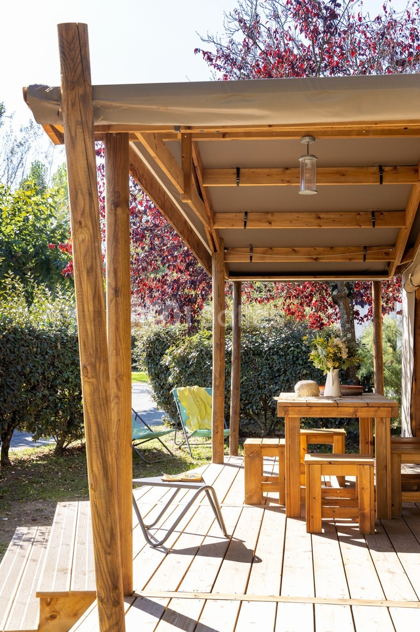 Terrasse en bois avec table, chaises et lanterne suspendue, entourée de végétation et éclairée par le soleil.
