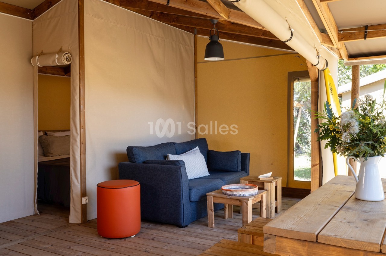 Intérieur d'une tente aménagée avec canapé, table basse, table en bois et bouquet de fleurs, éclairée par la lumière…