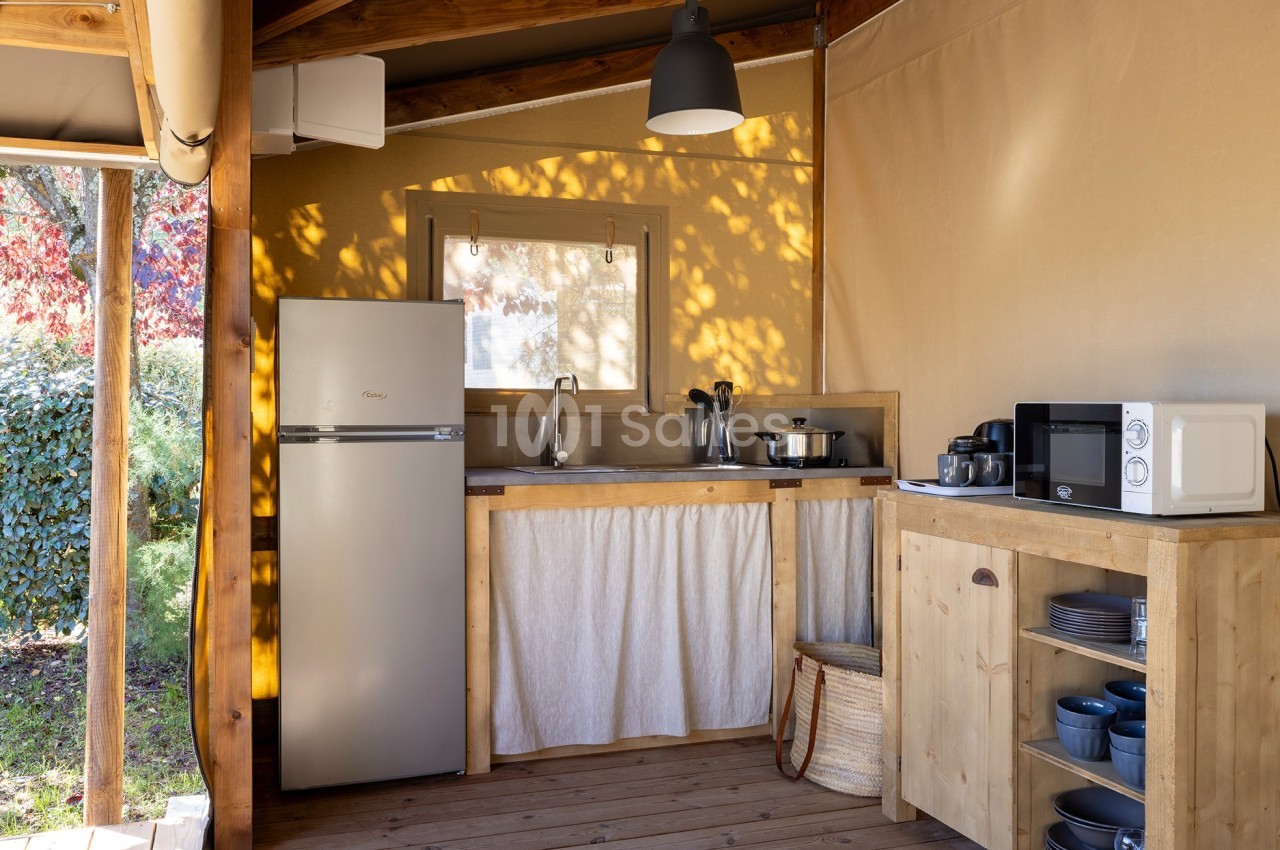 Cuisine extérieure en bois sous une tente, équipée d'un réfrigérateur, d'un évier, d'un micro-ondes et d'ustensiles.