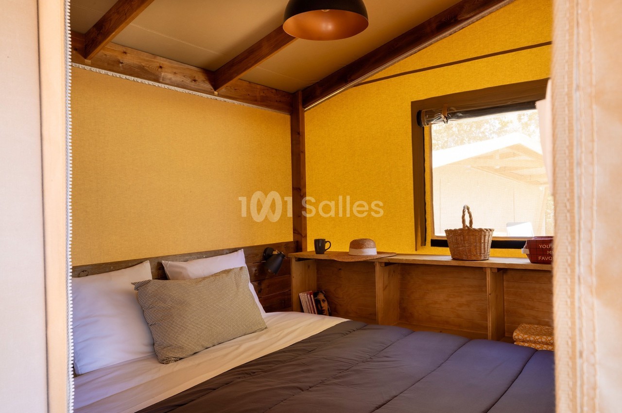 Intérieur d'une chambre en toile avec lit, coussins, étagère en bois, panier et fenêtre laissant entrer la lumière naturelle.