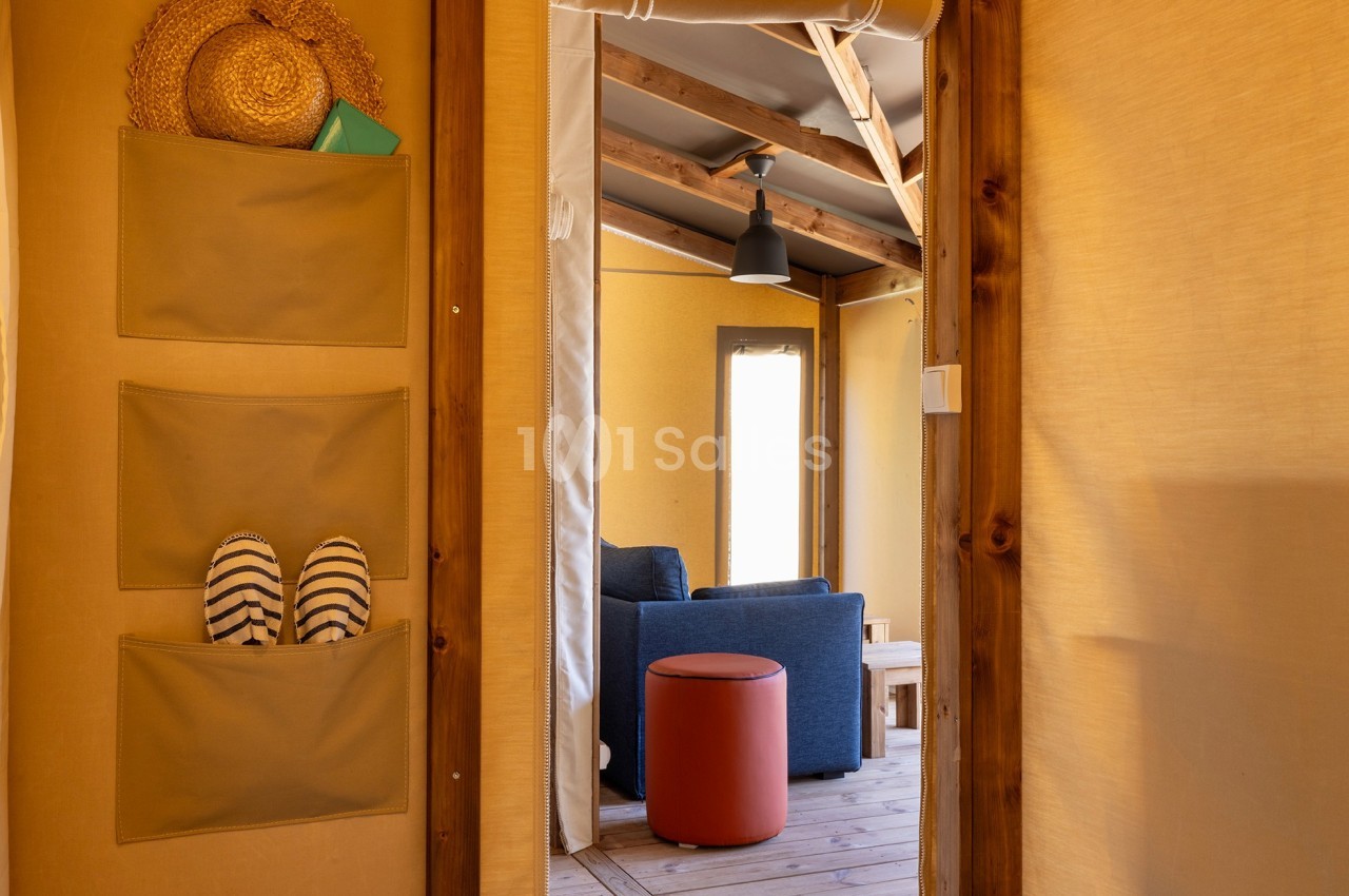 Vue intérieure d'une tente aménagée avec un salon, un tabouret rouge, des poches de rangement et des chaussures rayées.